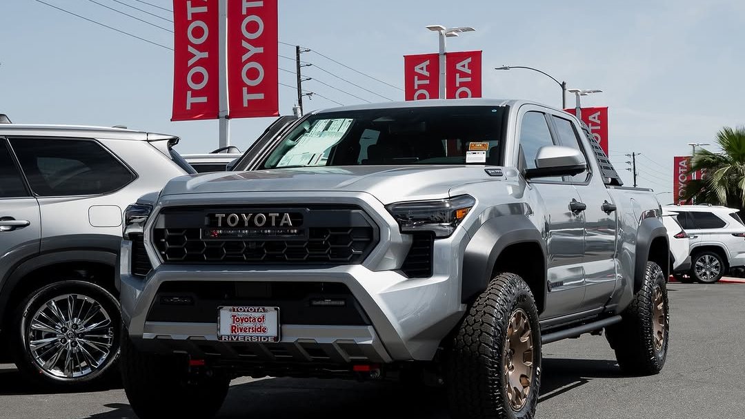 A silver 2026 Toyota Tacoma Trailhunter with bronze wheels on the Toyota of Riverside lot under red Toyota banners